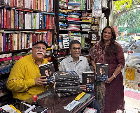 Anup Bahmi, DY Chandrachud and Mamta, in that order holding copies of Chandrachud's book Why Constitution Matters. Anup Banhmi and the former CJI are sitting behind the desk, while Mamta is standing, and there is are two book shelves in the background