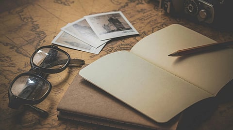 An open notebook and pencil rest on a vintage world map beside a pair of glasses and three black-and-white photos, evoking nostalgia and exploration.