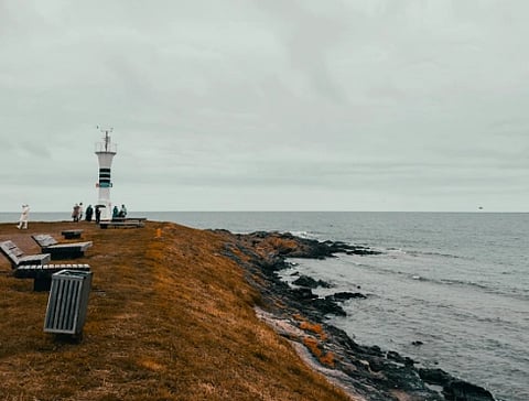 Coastal scene with a tall lighthouse and several people on a grassy shore. Overcast sky and benches line the path, leading to a calm sea. Tranquil mood.