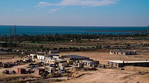 A serene coastal landscape with a cluster of small buildings and vehicles on sandy terrain. The sea and a distant tree line are visible under a clear sky.