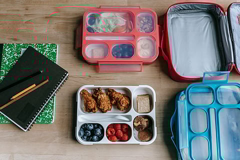 Organized lunch setup with fried chicken, berries, and snacks in divided containers. Nearby, notebooks with pencils on a wooden table.