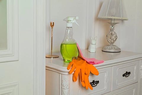 A spray bottle with cleaning liquid, a red sponge and orange rubber gloves placed on a white ornate cabinate in front of a white wall.