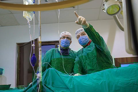 Two surgeons in green scrubs and masks focus intently as one points at a monitor. Medical equipment and an IV are visible, conveying a serious tone.
