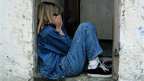A girl in a denim jacket and jeans sits in a doorway's corner. She covers her face with her hands, conveying sadness. The wall is worn and textured.