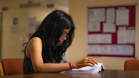 A woman with long dark hair and glasses is reading intently at a table in a quiet room. A bulletin board with papers is blurred in the background.