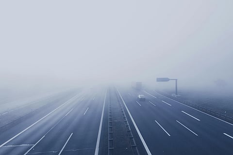Foggy highway view with sparse traffic, including cars and a truck, fading into dense fog. The mood is quiet and mysterious, emphasizing limited visibility.