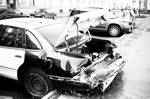 A black and white image shows a severely damaged car with a crumpled rear end parked on a street.