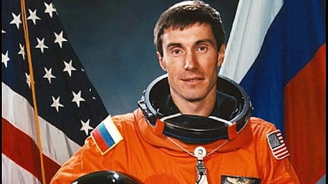 Cosmonaut Sergei Krikalev in an orange space suit holds a helmet, standing before American and Russian flags. His expression is composed and professional.