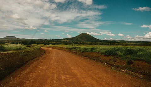 A red dirt road curves through a grassy landscape under a wide, blue sky with scattered clouds. Two green hills rise in the distance.