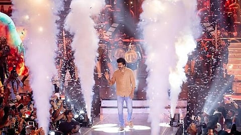 South actor Vijay on stage waves at a cheering crowd, surrounded by confetti and dramatic smoke effects.