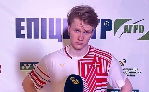 Anders Antonsen answering media questions, in his jersey, after a match, behind is a poster of events of Badminton tournaments and sponsors