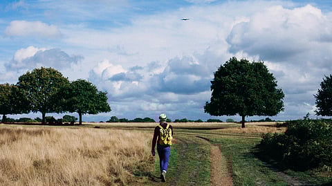  person with a yellow backpack walks along a dirt path through an open field under a blue sky with clouds, flanked by scattered trees.