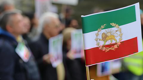 A hand holds the Iranian flag, amid a blurred crowd holding similar flags, conveying a sense of protest and unity.