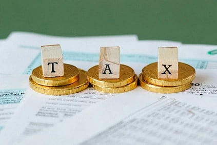 Three wooden blocks spelling "TAX" sit on stacks of gold coins atop tax documents, conveying financial responsibility and economic themes.