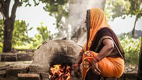 A woman in vibrant orange and red traditional attire tends to a wood-fired clay oven outdoors. Smoke rises amidst lush green trees