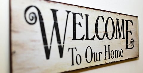 A rustic wooden sign with "Welcome To Our Home" in bold, decorative black lettering on a light background, conveying warmth and hospitality.