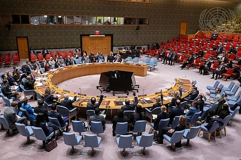 Spacious conference room with a large circular table; diverse group of people seated around it, engaged in discussion. Audience sits in red chairs.