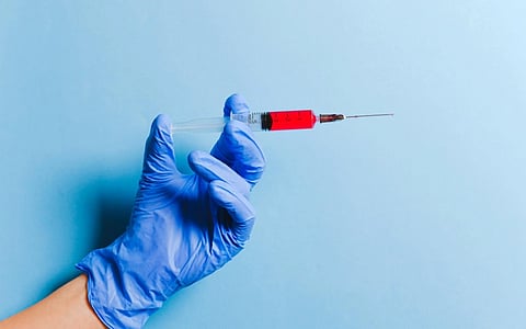 A hand in a blue glove holds a syringe filled with red liquid against a light blue background. The image conveys a clinical and sterile feel.