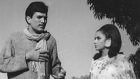 Actor Rajesh Khanna and actress Anju Mahendru standing by a lake in an outdoor setting. The man gestures while speaking, and the woman listens attentively. Both are wearing traditional Indian attire.