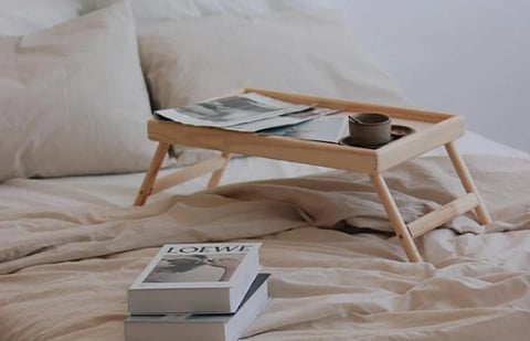 Cozy bed with beige sheets, fluffy pillows, and a wooden tray holding a cup and magazines. Nearby, a stack of books adds a relaxed, inviting vibe.