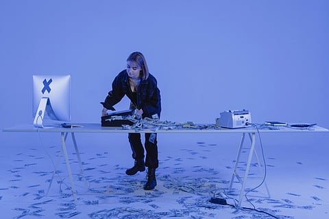 A person in dark clothing stands by a white table covered in scattered money and electronic devices, set in a blue-hued room creating a mysterious, high-tech ambiance.