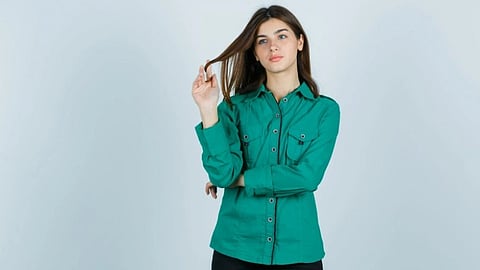 A woman in a green button-up shirt and black pants playfully twirls her hair. She stands against a light gray background with a calm expression.