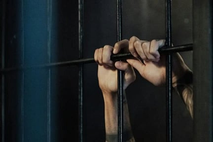 Hands grasping the bars of a dimly lit prison cell, conveying a sense of confinement and longing. The scene is somber and tense.