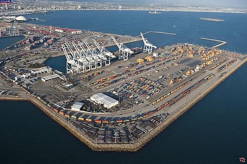Aerial view of a bustling container port, with large cranes, shipping containers, and cargo trucks. The surrounding calm sea contrasts the busy industrial scene.