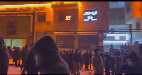 A crowd gathers on a city street at night, illuminated by warm orange building lights. People in dark clothing and hoodies convey a tense atmosphere.