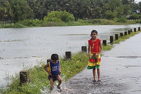 Children Playing in Rain Water on a road near a water body, with trees in the background.
