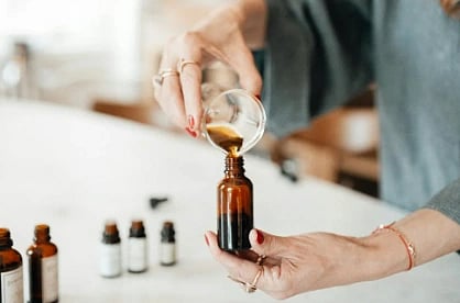 A woman pours liquid into a small brown bottle in a bright, softly focused kitchen. Nearby are several similar bottles, suggesting aromatherapy or skincare.