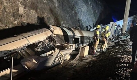 A derailed train lies on its side against a rocky cliff at night in Spain. Firefighters in yellow gear assess the scene, conveying urgency and caution.