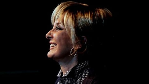 Googoosh with short blonde hair and a patterned blouse smiles off-camera against a dark background, conveying a sense of joy and confidence.