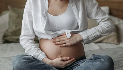A pregnant woman sits cross-legged on a bed, gently cradling her belly. She's wearing a white shirt and jeans. The mood is calm and nurturing.
