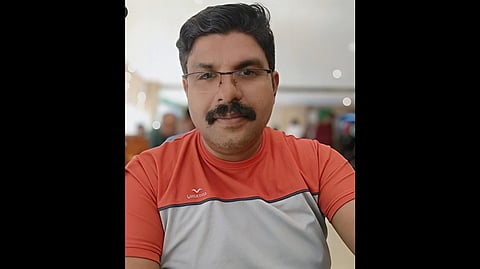 42-year-old Kerala man U Deepak with glasses and a mustache, wearing a red and gray sports shirt, smiles at the camera. The background is blurred, suggesting an indoor setting.