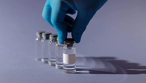 A blue-gloved hand picks up one of several aligned clear vials on a smooth, gray surface, conveying a clinical and precise atmosphere.