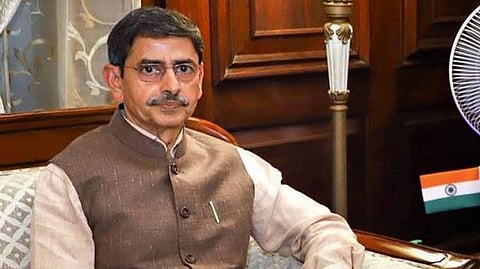 Tamil Nadu Governor R N Ravi in a brown vest sits on a sofa in an ornate room, holding a book. An Indian flag is visible in the background