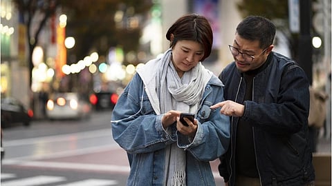 A woman and a man stand on a city street in the evening, focused on a smartphone. The man points at the screen, suggesting engagement and curiosity.