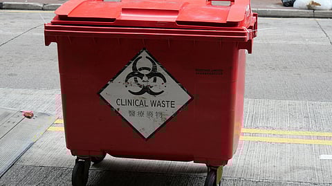 A red clinical waste bin with biohazard symbol and warning text, on a street, conveying caution. People walk by in the background, adding urban context.