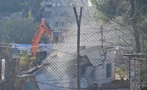 A wired fence, behind it a UNRWA building, a crane destroying the building, dust flying all around due to the demolition, trees in the background