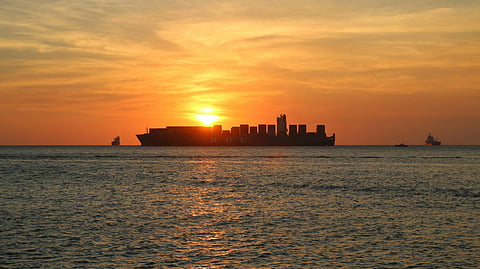 Shipping vessel silhouetted against a vibrant sunset over calm ocean. Warm tones of orange and yellow fill the sky