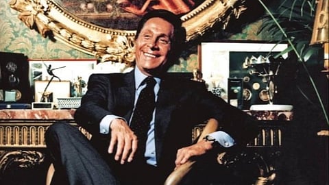 The legendary Italian fashion designer Valentino Garavani dressed in a suit, sits confidently on a horned chair in a lavish room, surrounded by art and trophies. The ambiance is luxurious and sophisticated.