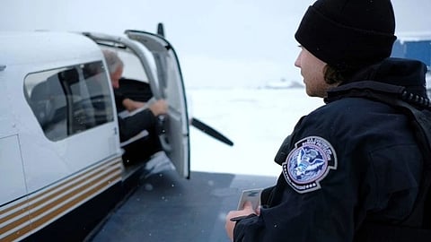 A security officer in a dark uniform and wool hat observes a person entering a small aircraft on a snowy day. The mood is vigilant and focused.