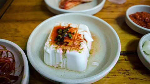 A white block of tofu with red sauce and chopped green onions on a plate. Surrounding dishes contain kimchi and sauces, set on a wooden table.