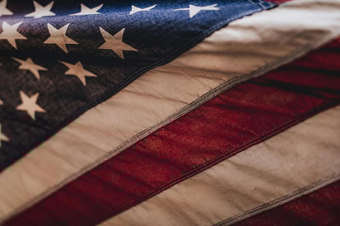 Close-up of a textured American flag, showing its red and white stripes and white stars on a blue field. The image conveys a sense of patriotism.
