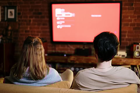 A couple sits on a sofa in a cozy living room with brick walls, facing a large red TV screen displaying a menu. 