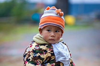 A small child dressed warmly in a colorful jacket and orange-striped hat stands on a dirt path. The child looks curiously at the camera.