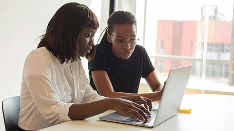 Two people work together at a laptop in a bright room. One points at the screen while the other listens intently, conveying teamwork and focus.