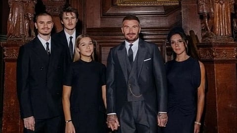 The formally dressed Beckham family of five poses indoors against a wooden backdrop adorned with sculptures.
