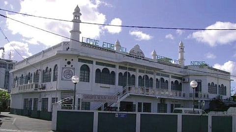 Two-story white mosque with a large green sign reading "Love for All, Hatred for None" on the roof. The building features multiple arches and minarets, set against a clear blue sky.